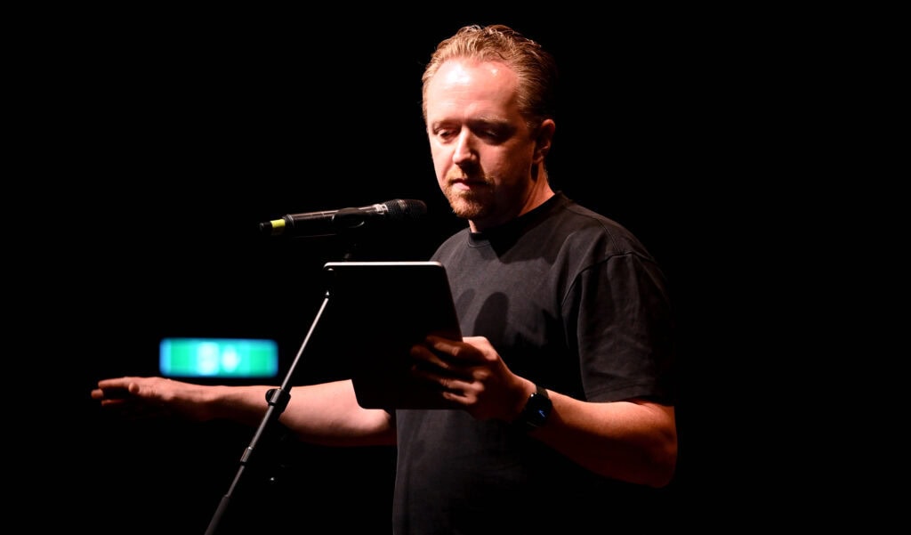 Freier Redner hält eine Rede auf einer Bühne, liest aus einem Tablet, im Hintergrund dunkle Beleuchtung, Fokus auf die Präsentation.