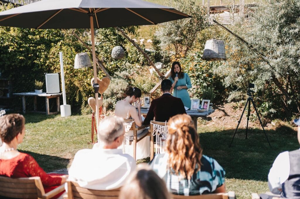 Freie Redner hält eine Trauung im Garten unter einem Sonnenschirm, umgeben von Natur, mit Gästen, die aufmerksam zuhören. Perfekt für Hochzeiten im Freien.