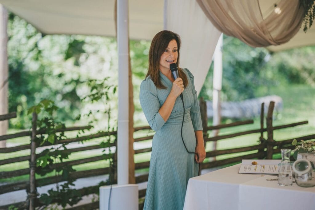 Frau hält Mikrofon bei einer Rede im Freien, professionelle Rednerin bei Event, natürliche Umgebung, Fokus auf Präsentation, Event-Redner, öffentliche Ansprache, Outdoor-Veranstaltung.