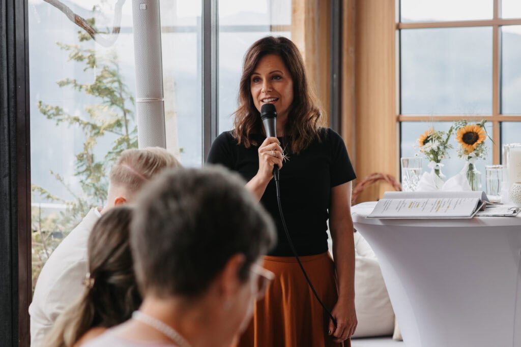 Frau hält Mikrofon bei Vortrag, umgeben von Zuhörern, in hellem, modernem Raum mit großen Fenstern und Sonnenblumen.