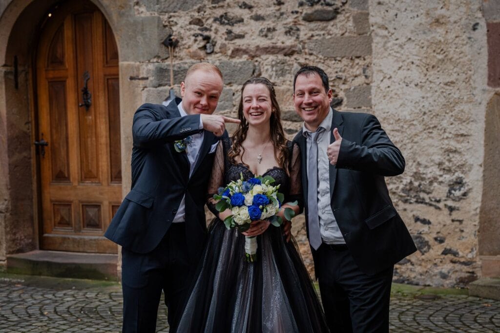 Hochzeitspaar mit Redner bei einer Zeremonie im Freien, lachend und glücklich, umgeben von historischem Steinmauer und Holztür, ideal für Hochzeitsreden und Feierlichkeiten.