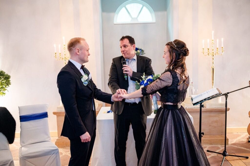 Ein Redner hält eine Hochzeitsrede bei einer standesamtlichen Trauung, während das Brautpaar sich die Hand reicht. Die Zeremonie findet in einem eleganten Raum mit Kerzen und festlicher Atmosphäre sta.