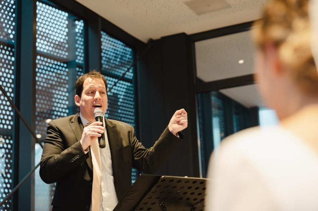 Motiv eines Redners, der bei einer Veranstaltung spricht, mit Mikrofon in der Hand, vor Publikum, in einem modernen Raum mit großen Fenstern.