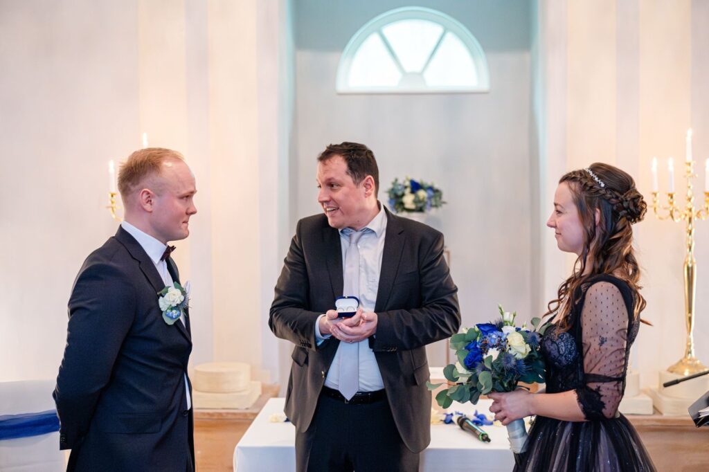 Hochzeitsredner hält eine Rede bei einer standesamtlichen Trauung mit einem Brautpaar und einem Zeremonienmeister in einem eleganten Raum.