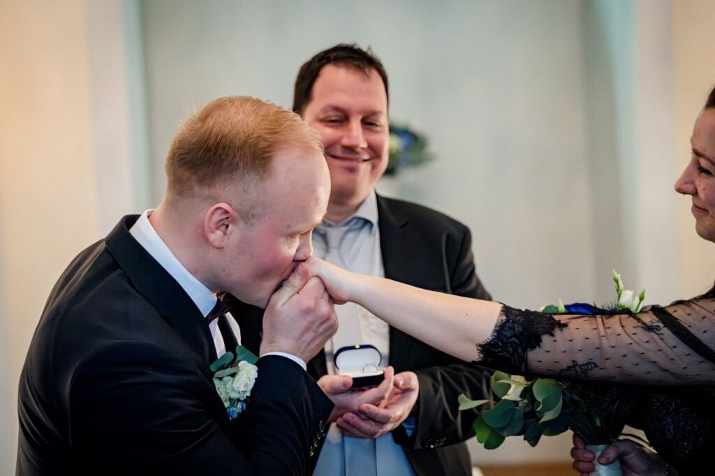 Ein Mann wird während einer Hochzeitszeremonie von einer Frau einen Ring auf den Finger gesteckt, während ein Trauredner die Zeremonie leitet.