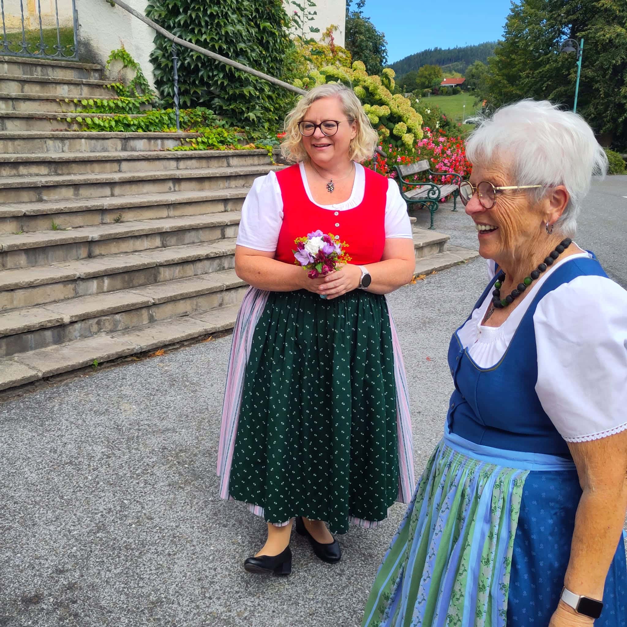 Zwei Frauen in traditionellen Trachten bei einer Freie Redner Zeremonie im Freien, lachend und mit Blumen, umgeben von Natur und Blumen, bei schönem Wetter.