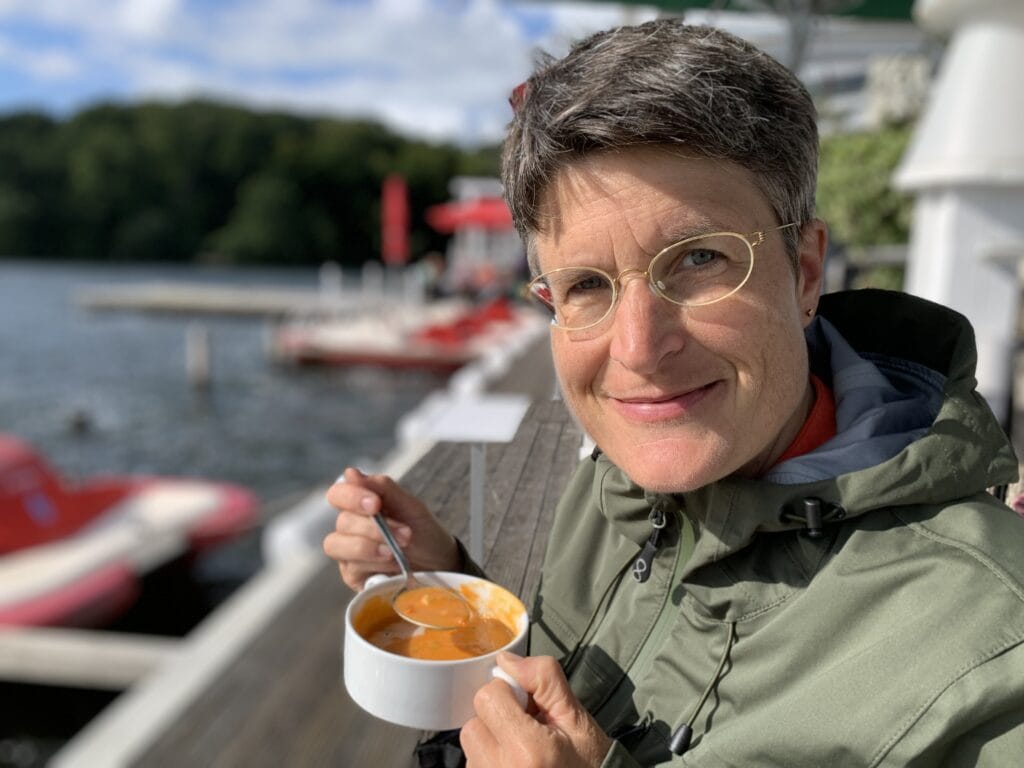 Freundlicher Redner mit Brille hält eine Schüssel mit Suppe am Wasser, im Hintergrund Boote und Natur, ideal für Redner-Events.