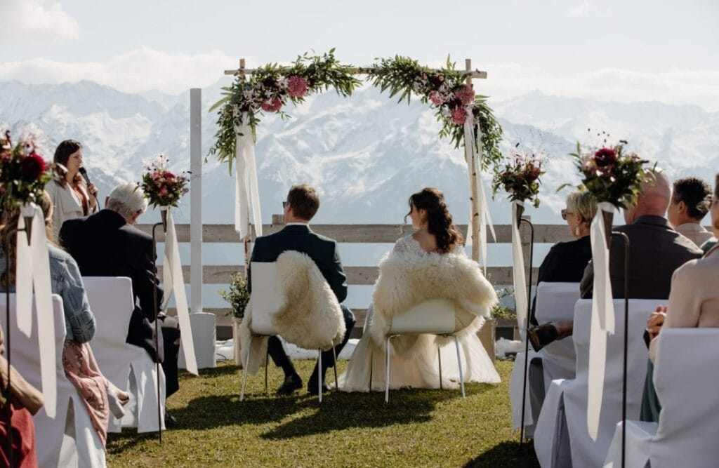 Romantische Hochzeit im Freien mit Bergpanorama, Braut und Bräutigam unter einem Blumenbogen, umgeben von Gästen, bei schönem Wetter.