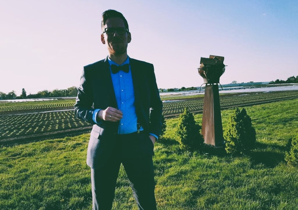 Ein freier Redner steht in einem grünen Feld bei Sonnenuntergang, trägt einen schwarzen Anzug mit blauer Krawatte und hält eine Hand in der Tasche. Perfekt für Hochzeiten, Trauungen und Events.