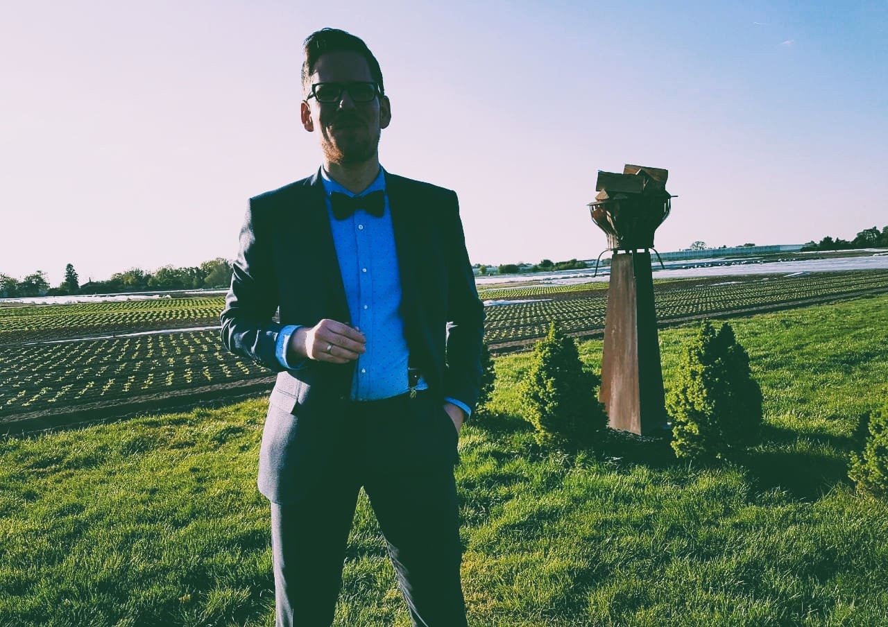 Freier Redner Michael Kniess steht in einem grünen Feld bei Sonnenuntergang, trägt einen schwarzen Anzug mit blauer Krawatte und hält eine Hand in der Tasche. Perfekt für Hochzeiten, Trauungen und Events.