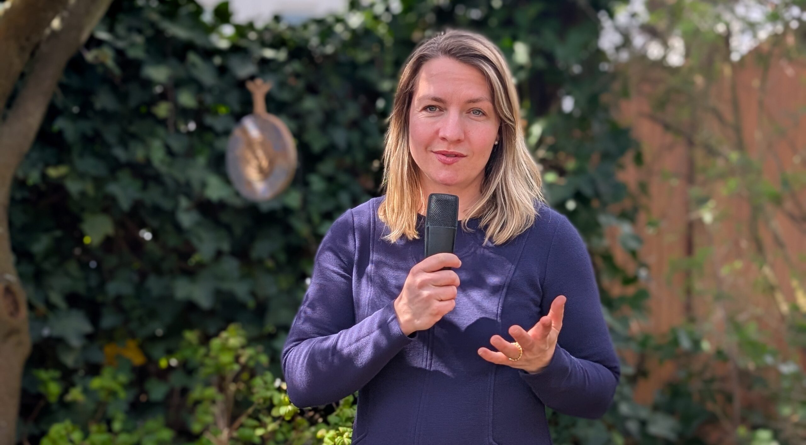 Traurednerin Kathi Kaufmann hält eine Rede im Garten, spricht in ein Mikrofon, umgeben von grünen Bäumen und Pflanzen, ideal für professionelle Redner und Moderatoren bei Veranstaltungen.