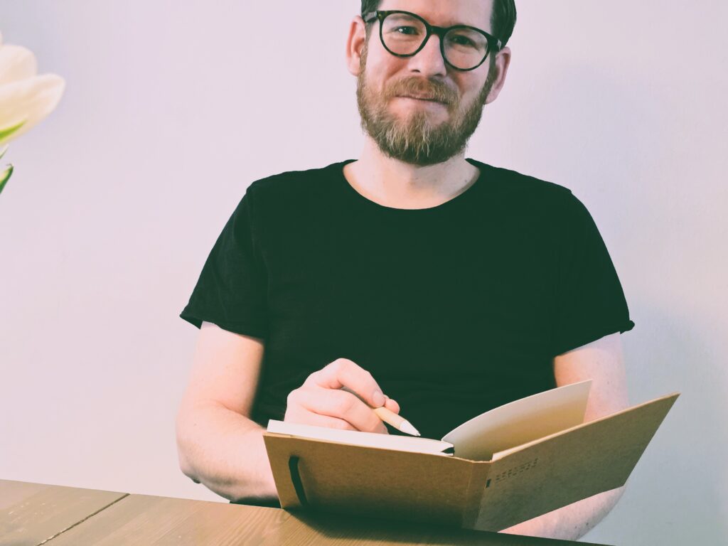 Ein freundlicher Redner mit Brille und Bart, der bei einer Veranstaltung spricht, umgeben von einer professionellen Atmosphäre.