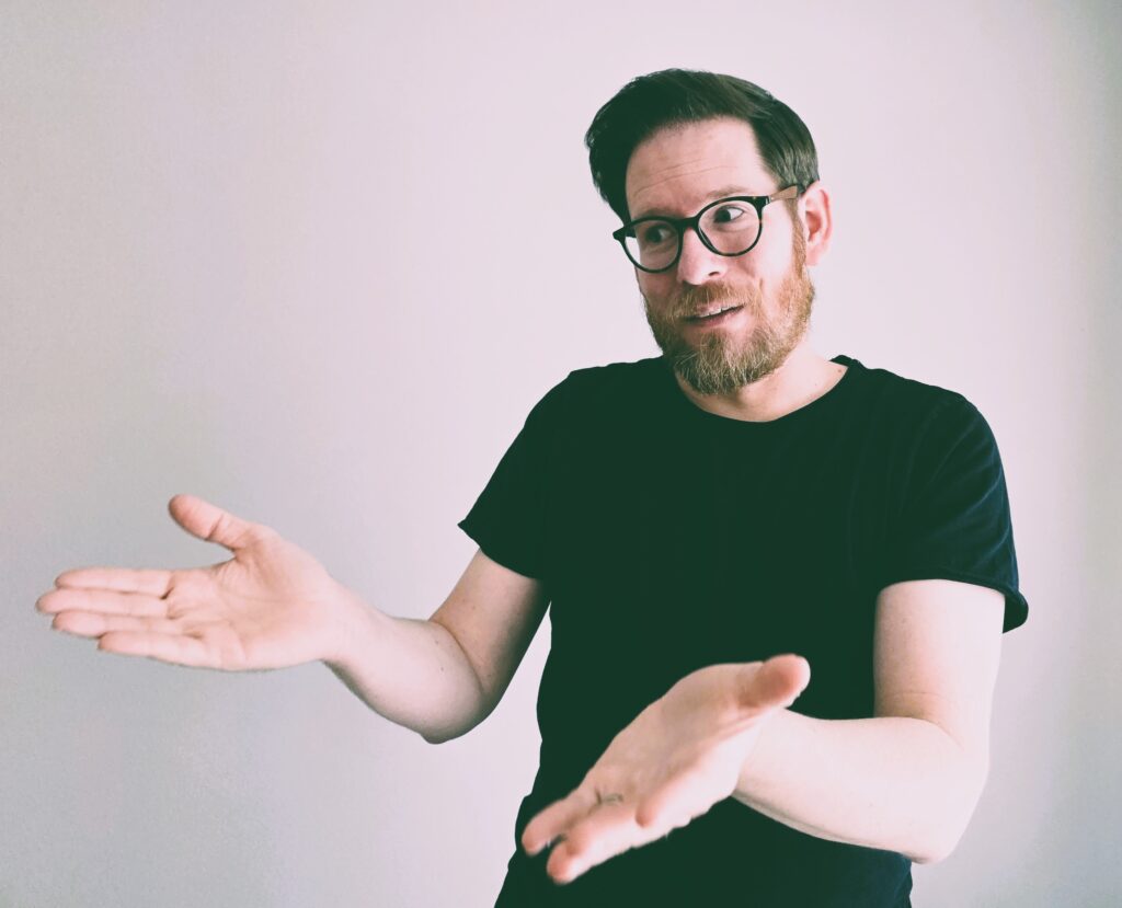 Mann mit Brille und dunklem T-Shirt, der eine Geste macht, symbolisiert Redner bei einer Veranstaltung. Perfekt für professionelle Redner, Moderation und Eventplanung.