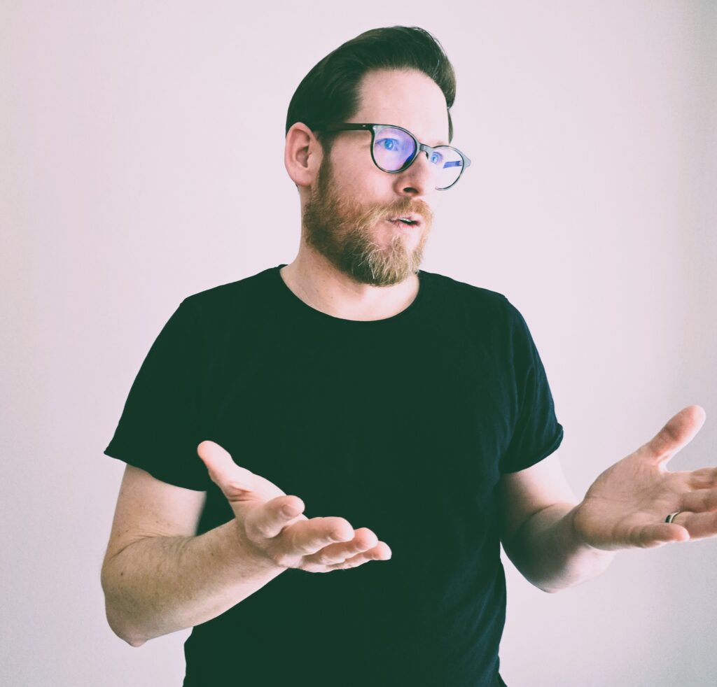 Mann mit Brille und schwarzem T-Shirt spricht offen, symbolisiert professionelle Redner für Veranstaltungen und Vorträge.