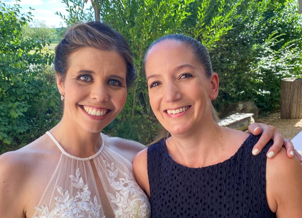 Zwei Frauen bei einer Hochzeit im Freien, eine trägt ein weißes Brautkleid, die andere ein dunkles Kleid, umgeben von grüner Natur, symbolisieren freie Redner für persönliche Hochzeitsreden.