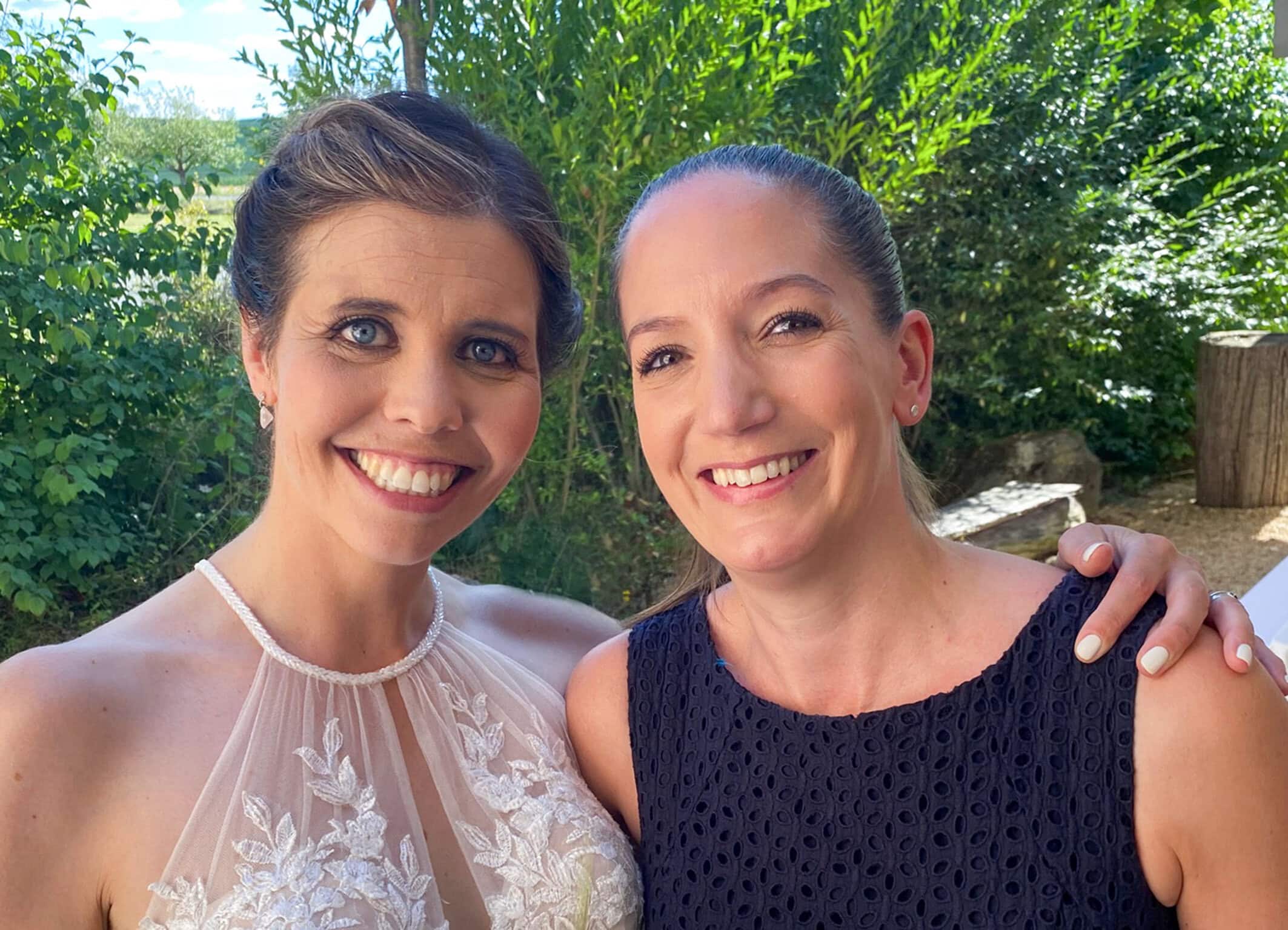 Zwei Frauen bei einer Hochzeit im Freien, eine trägt ein weißes Brautkleid, die andere ein dunkles Kleid, umgeben von grüner Natur, symbolisieren freie Redner für persönliche Hochzeitsreden.