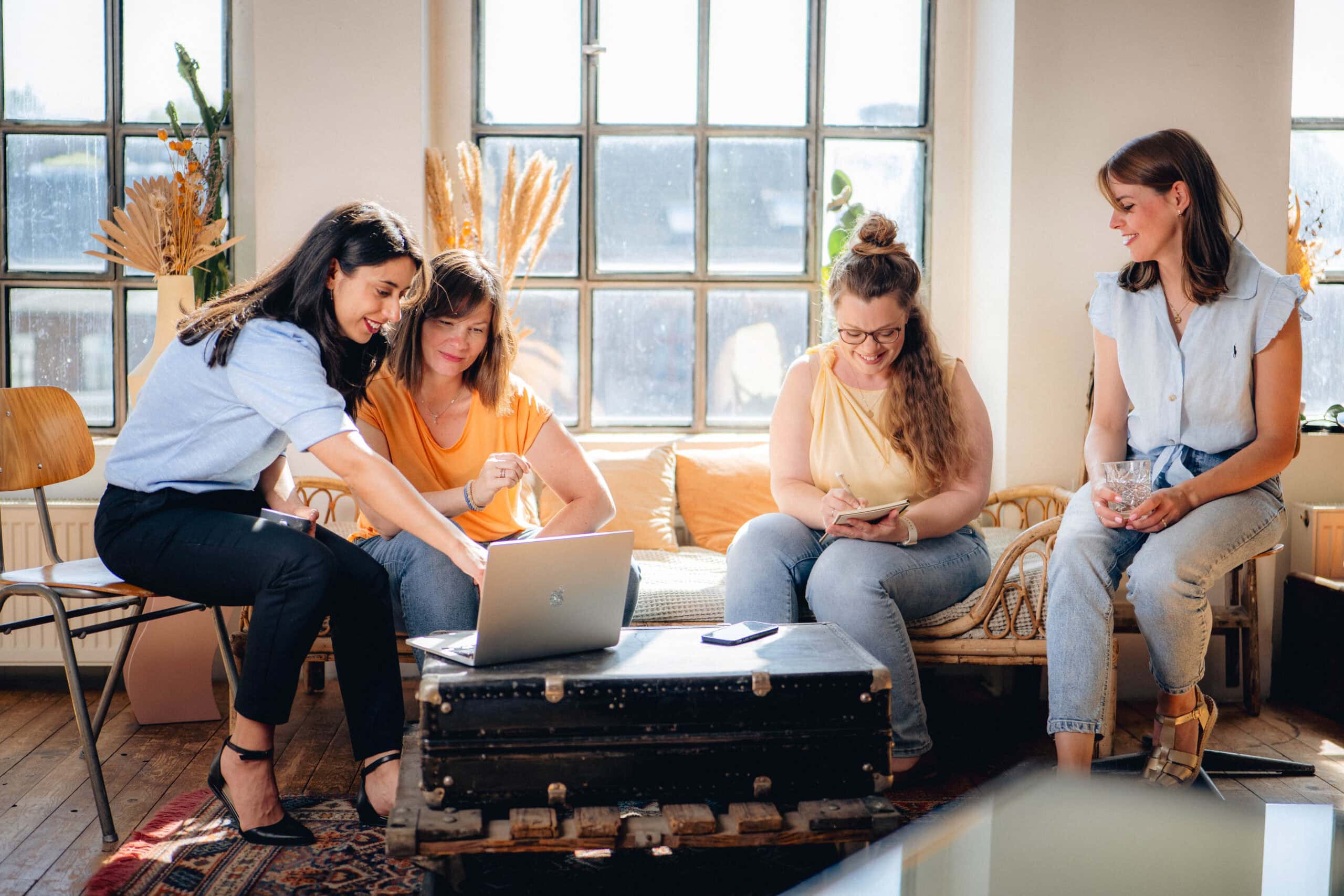 Vier Frauen sitzen in einem hellen Raum, arbeiten gemeinsam an einem Laptop und unterhalten sich, umgeben von natürlichen Lichtquellen und dekorativen Pflanzen.