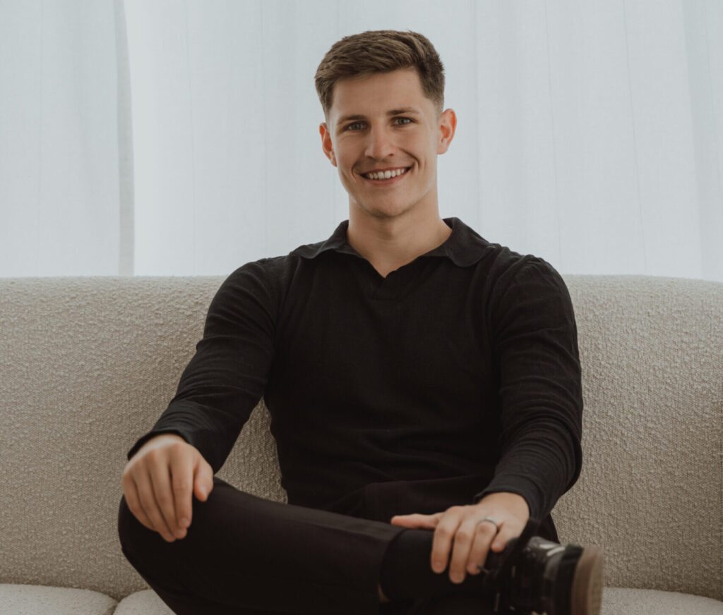 Junger freier Redner sitzt entspannt auf einem Sofa, lächelt freundlich und wirkt professionell für öffentliche Reden und Präsentationen.