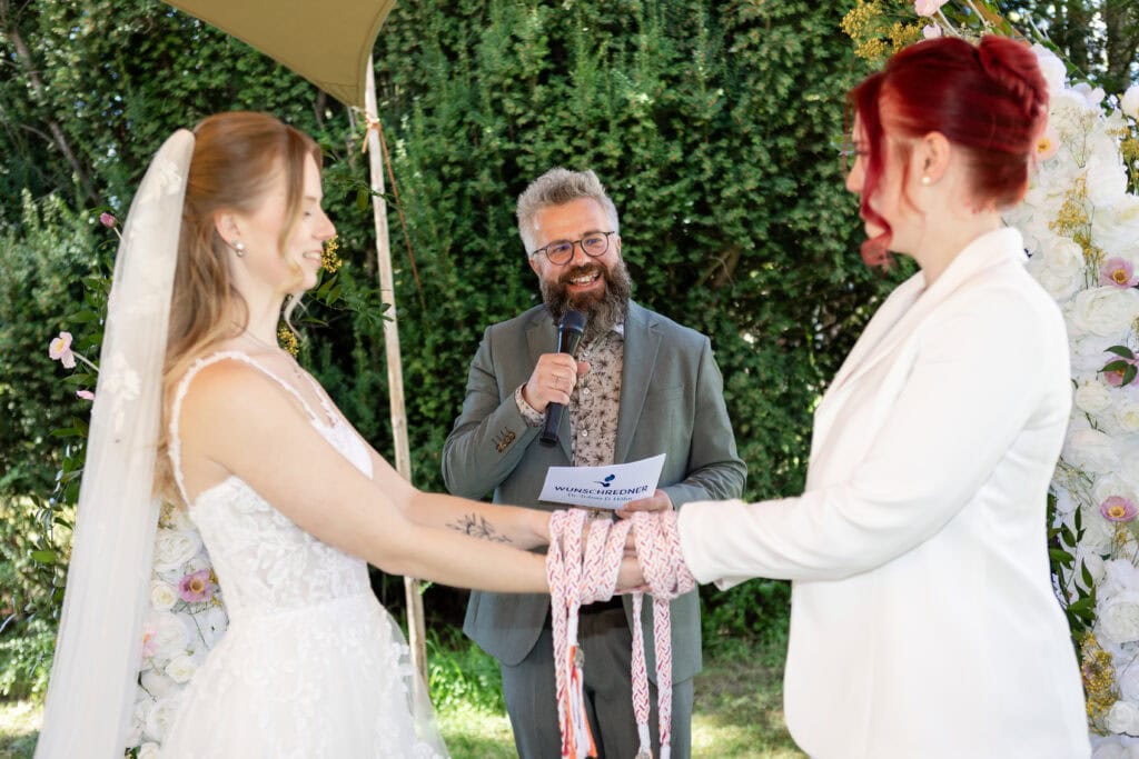 Hochzeit im Garten mit freiem Redner, Braut und Brautmutter beim Austausch der Ringe, fröhliche Atmosphäre, grüne Natur im Hintergrund.