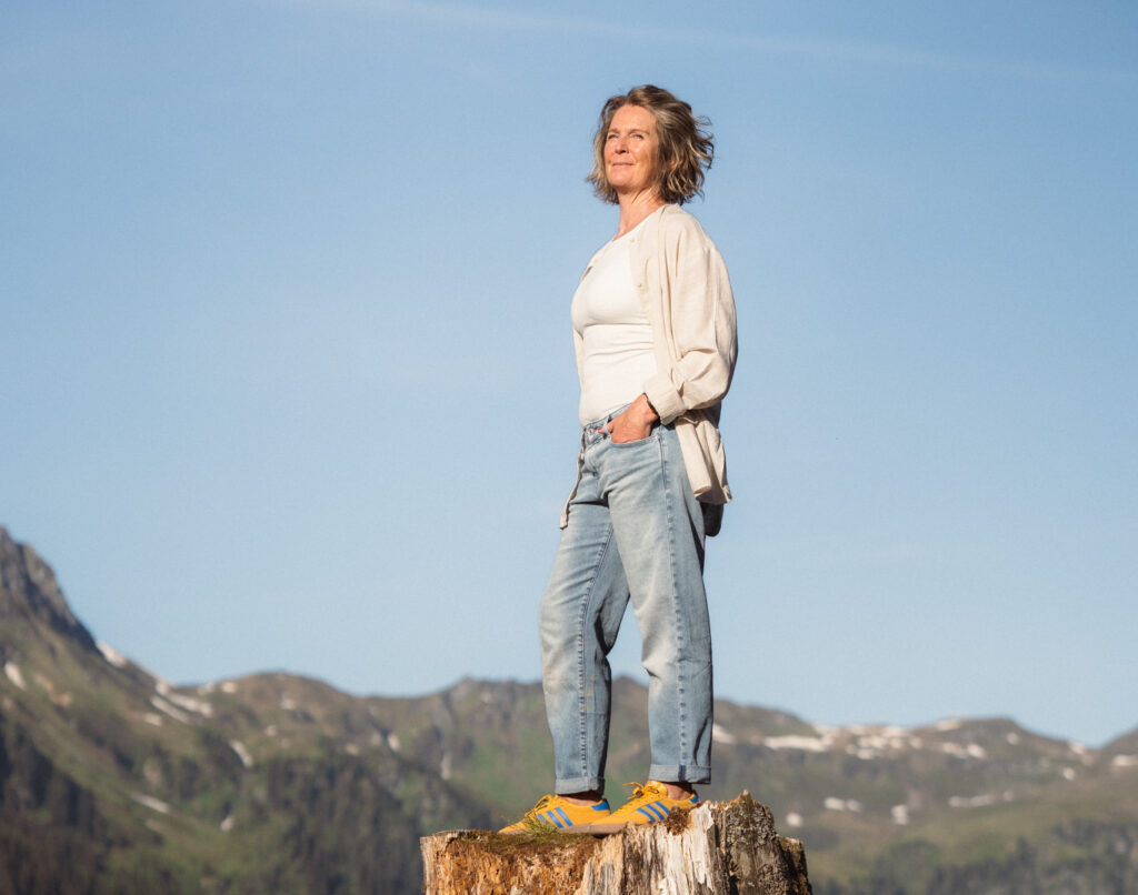 Freie Rednerin steht auf einem Baumstumpf in der Natur mit Bergen im Hintergrund, symbolisiert Selbstvertrauen und Freiheit.