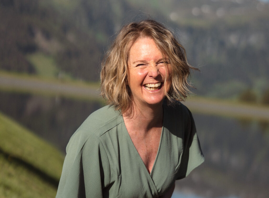 Freie Rednerin lachend im Freien vor Berglandschaft und See, natürliche Umgebung, positive Stimmung, authentisch, professionell, für Redner-Website optimiert.