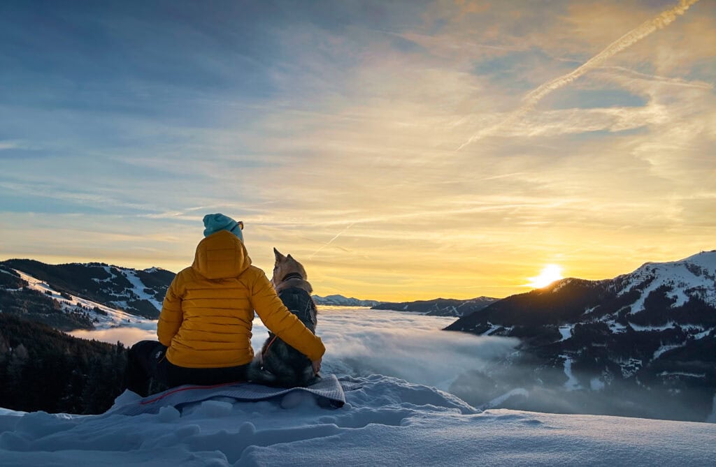 Person und Hund sitzen auf Schneedecke und genießen den Sonnenuntergang in den Bergen bei winterlicher Landschaft.
