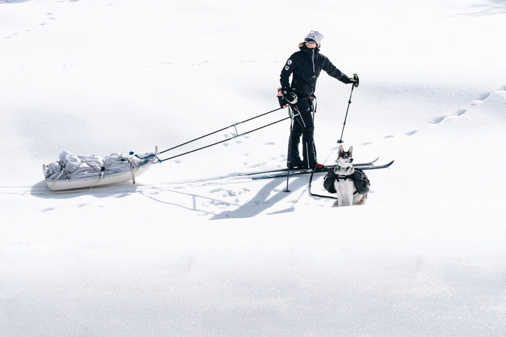 Skifahrer mit Hund in verschneiter Landschaft, zieht einen Schlitten mit Gepäck.