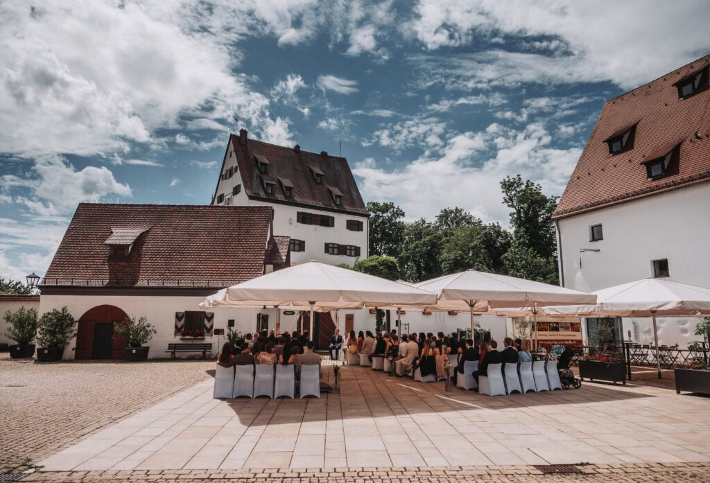 Freie Redner bei einer Outdoor-Veranstaltung im historischen Dorf mit weißen Zelten und Gästen unter einem blauen Himmel.