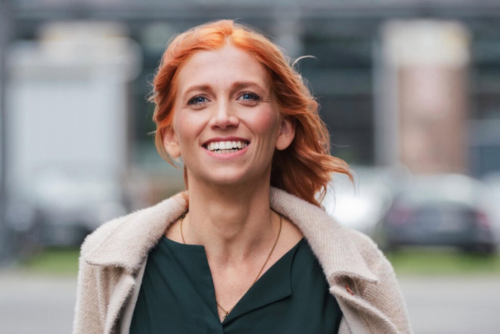 Freie Redner Frau mit roten Haaren, lächelnd, im urbanen Umfeld, professionelle Rednerin für Veranstaltungen und Anlässe.